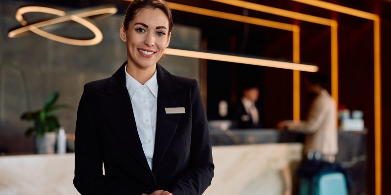 Smiling,Female,Hotel,Receptionist,In,A,Hallway,Looking,At,Camera.